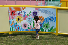 写真：壁に取り付けられた幅180センチほどのパネルとその前に立つ女の子。パネルに描かれた8つの花にはそれぞれドレミファソラシドの文字と、その上に「花のうえに手をのせると音が出るよ！」の説明