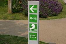 写真：園路の分かれ道にある道標。緑のパネルに白抜きの字。大きな矢印や勾配が急になることを示すピクトグラムも示されている