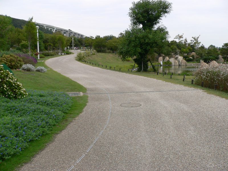 写真：緑の庭園の間を延びる幅の広い園路。点と線が連続した、視覚障害者誘導用パイプが1本延びている