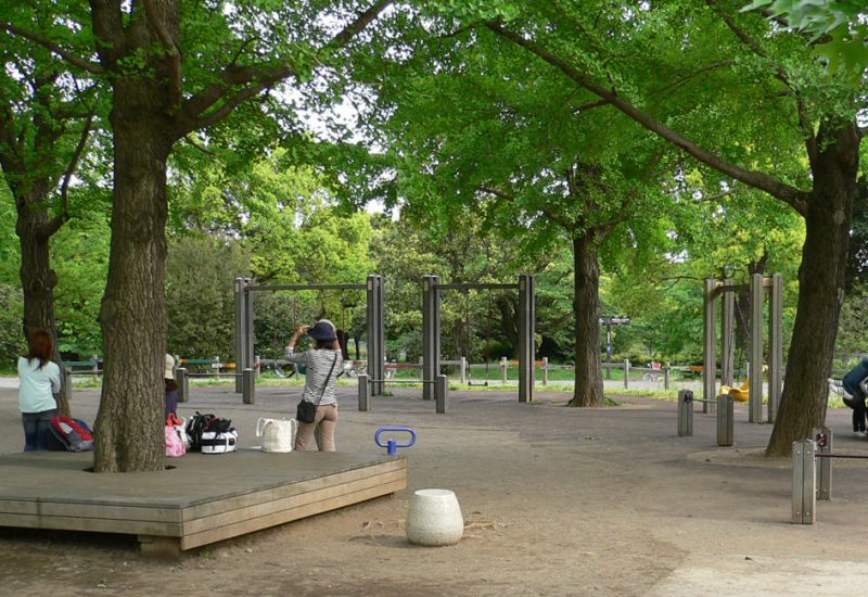 写真：遊び場のあちこちに、緑豊かな枝を大きく広げた木々。木の根元に作られた大型のベンチに子どものためのたくさんの荷物を置いて談笑するお母さんたち