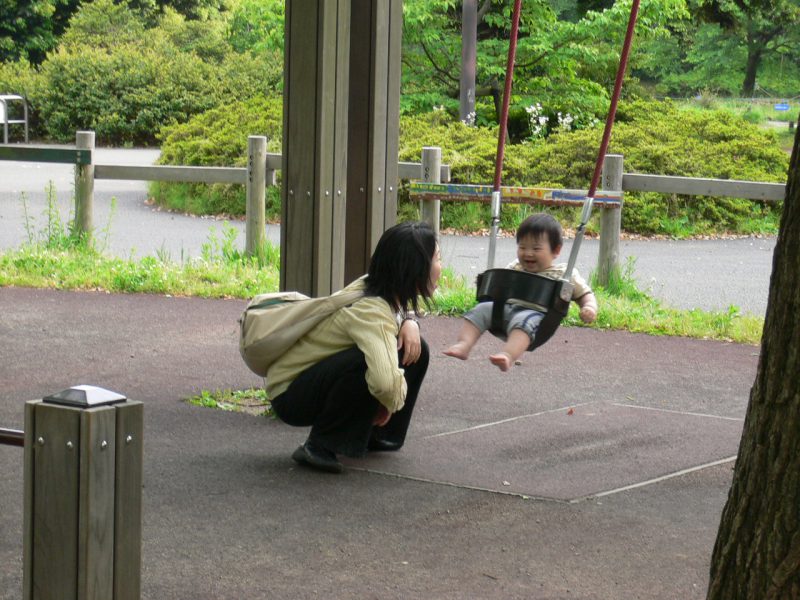 写真：幼児用のブランコに乗りご機嫌のあかちゃん。地面にしゃがんであかちゃんと笑顔を交わすお母さん