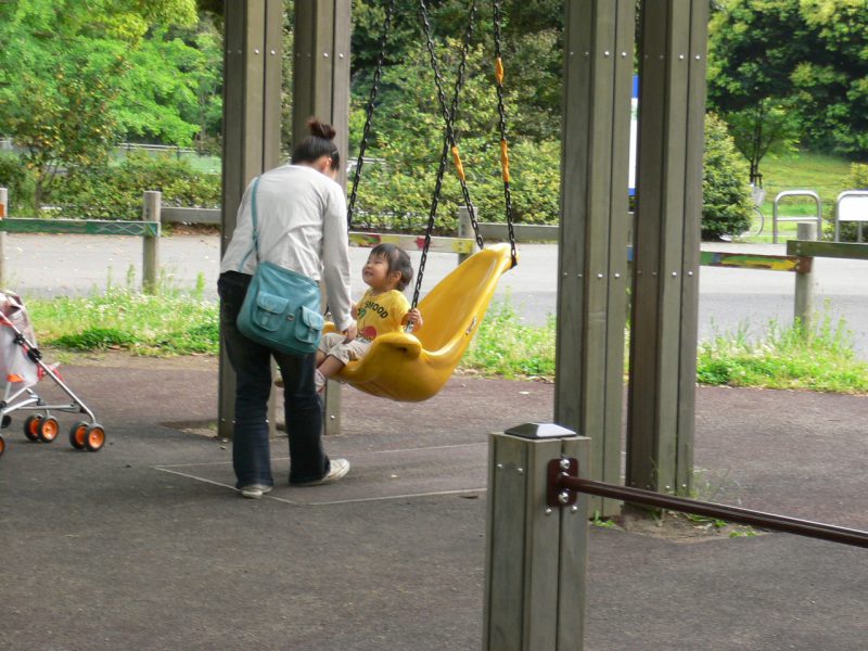 写真：背もたれ付きブランコに乗って笑顔になった小さな女の子。お母さんが正面に立ちブランコをそっと押す