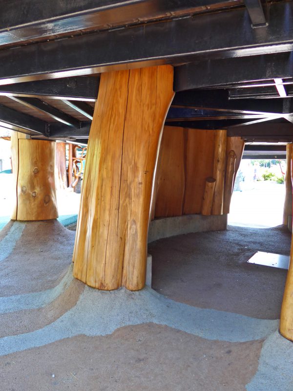 写真：デッキ下は天井高が低く、木の柱が立ち並ぶ。地面には茶色とグレーのゴムチップ舗装が用いられ、柱の根元部分が盛り上がっている