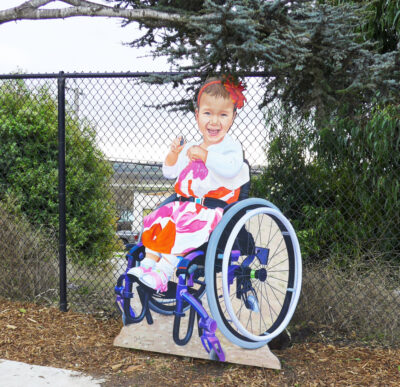 写真：遊び場にあるテイタムの肖像パネル。髪飾りを付け笑顔で車いすに乗る3才の女の子