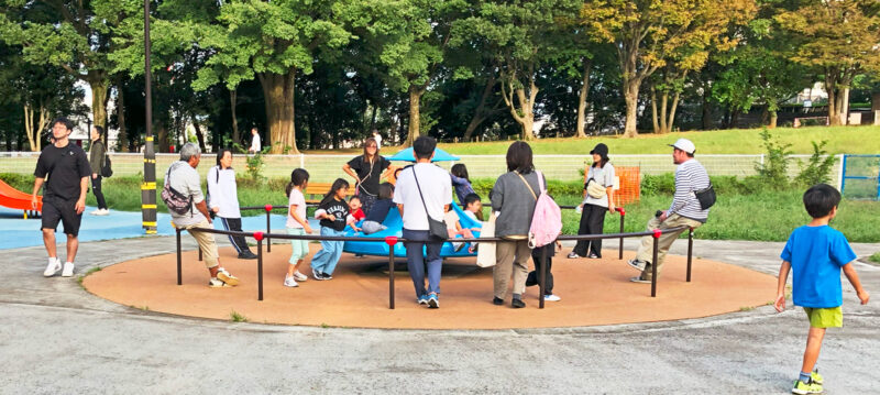 写真：回転遊具で遊ぶ様々な年齢の子どもたちと、安全柵に腰掛けてにこやかに見守る大人たち。