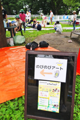 写真：出入口の正面に置かれたイベントの案内板。草地には持ち物を置いたり子どもが寝転んだりできるシートが敷かれている。