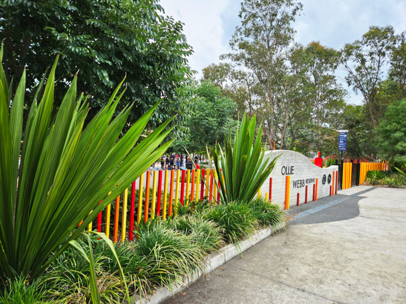 写真：色とりどりの柵と大小の植物が並んだエントランス周辺。奥には公園名が大きな凸文字で記された石塀。