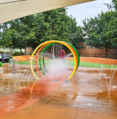 写真：直径約2メートルのトンネルをキックボードで行く男の子。自動洗車機を通り抜ける車のよう。周囲の地面からも様々な角度で水が噴き出している。