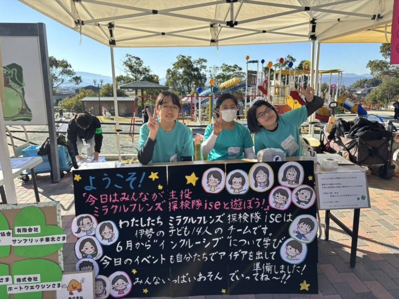 写真:受付テントで笑顔の子どもメンバー3人。前には歓迎&チーム紹介のメッセージが書かれた看板。