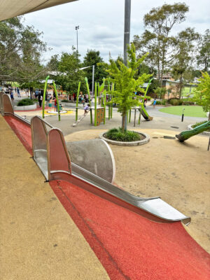 写真：ステンレス製で幅広の滑り台が2本。その間に大きな土管のようなトンネルがある。