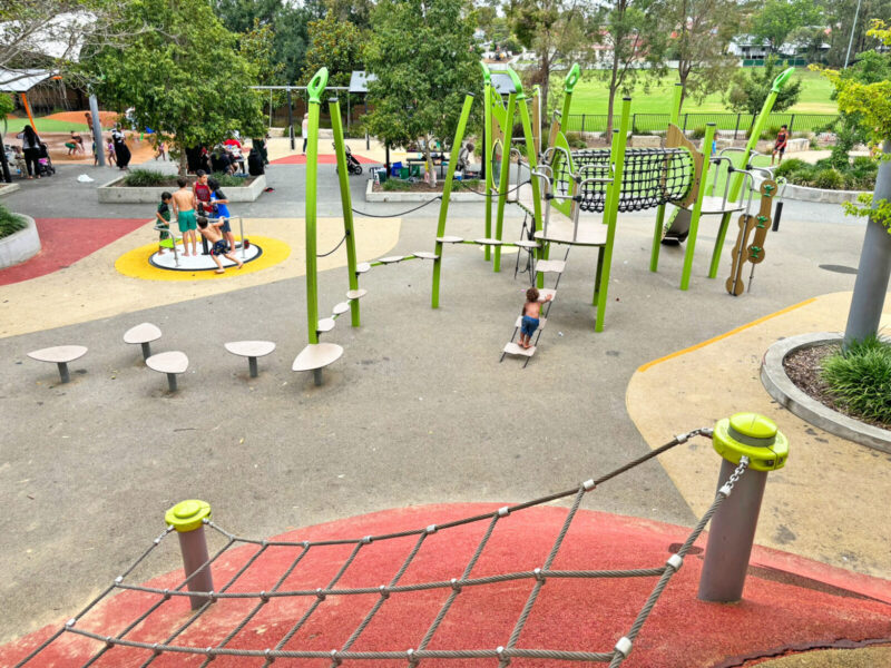 写真：頂上から見下ろす回転遊具や小さな複合遊具、スプラッシュパッドエリア。子どもたちが水着や裸足で遊んでいる。