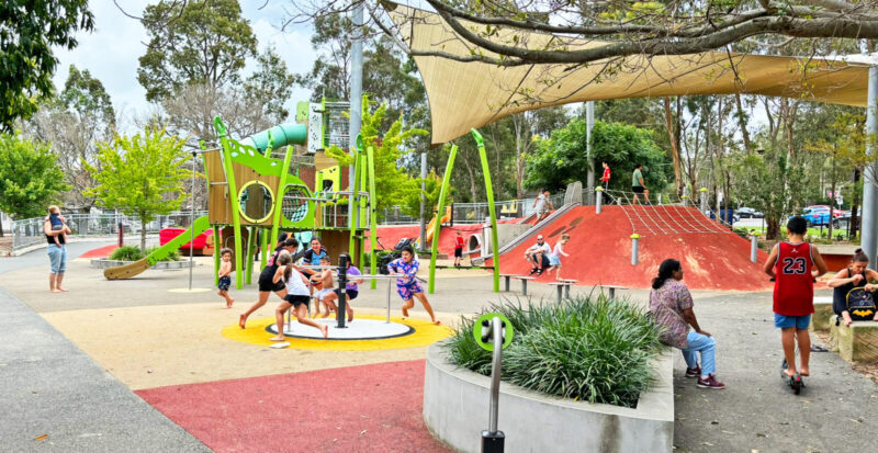 写真：遊び場の遠景。回転遊具や土手で遊ぶ子どもたちや園路をキックボードで進む男の子。ベンチなどでくつろいで見守る大人たち。